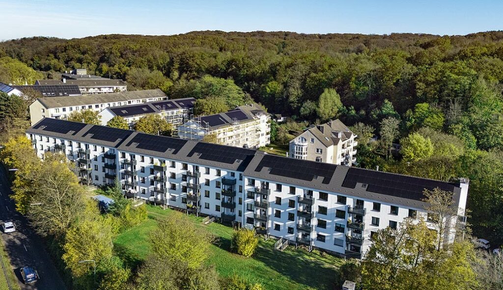 Luftaufnahme einer Siedlung mit Solardächern, Wald im Hintergrund, sonniger Tag, blauer Himmel
