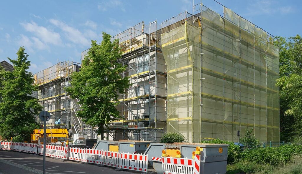 Eingerüstetes Haus, 4-geschossig, davor rot-weiße Absperrungen vor Bauschutt-Containern, blauer Himmel